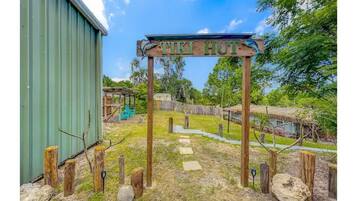 Une enseigne en bois portant l’inscription « TIKI HUT », placée dans un décor extérieur rustique, avec un bâtiment vert, des poteaux en bois et une structure à toit de chaume.