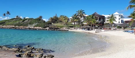 A beach with clear turquoise water, white sand, and palm trees. There are buildings with a modern design in the background.