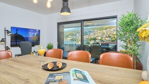 A modern dining area with a wooden table, leather chairs, a city view through large windows, and a television on the wall. - Hedera Estate, Hedera A15 (Dubrovnik)