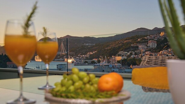 A table with two glasses of a yellowish drink, a plate of green grapes, and an orange, with a view of a city and mountains in the background. - Hedera Estate, Hedera A15 (Dubrovnik)