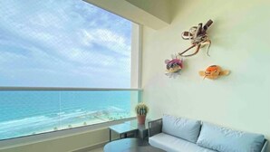 A modern living room with a large window overlooking the ocean, a light blue sofa, a small round table, and decorative wall art. - 🏝🏝Best View, Pool, Jacuzzi, Gym, WiFi, BBQ, Parking 🏝🏝 (Playa Juan Dolio)