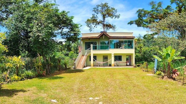 A two-story house with a balcony, surrounded by lush greenery and a clear sky. - Pacífico Suítes (Ubatuba)