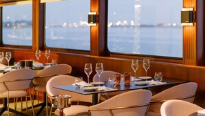 Un wagon-restaurant avec des tables pour deux personnes, équipé de verres à vin et de couverts, et de grandes fenêtres offrant une vue sur l’eau et les lumières de la ville.