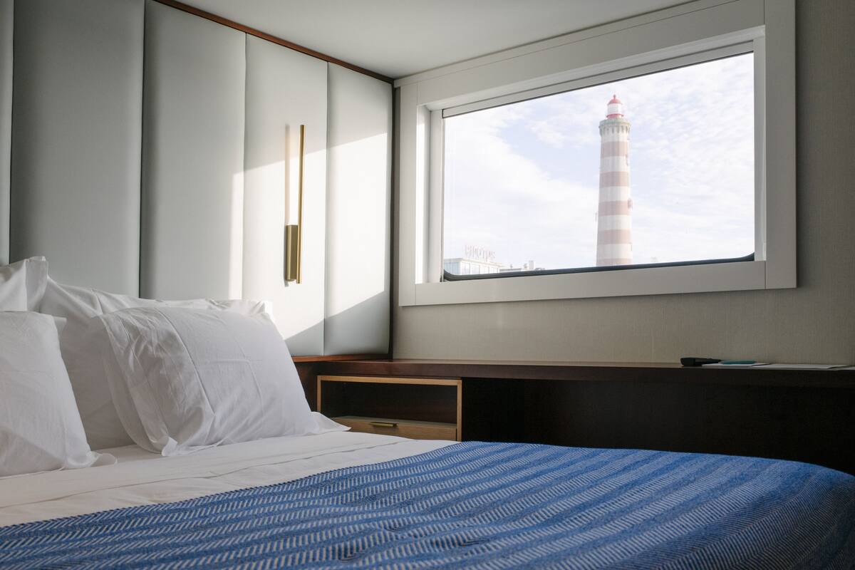 Habitación de hotel con cama, mesita de noche, ventana con vista a un faro y una lámpara fijada en la pared.