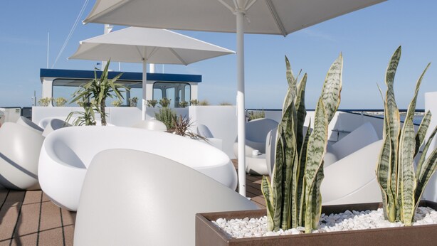 Une terrasse sur le toit, dotée de chaises longues blanches au design moderne, d’un grand parasol et d’un jardinier contenant des plantes à rayures.