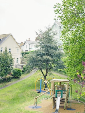 A playground with a slide and swings, surrounded by grass and trees, with houses in the background.