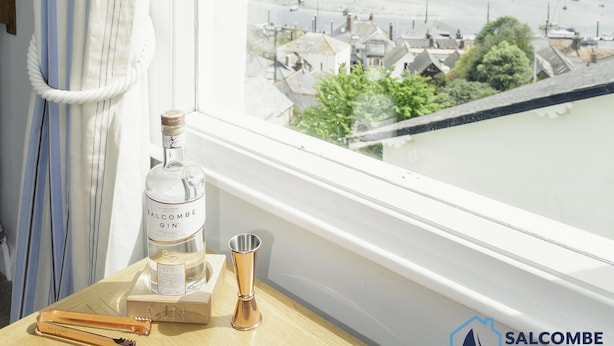 Una botella de ginebra Salcombe Abode, un shaker de cócteles y un destornillador de corcho sobre una mesa de madera junto a una ventana con vistas al puerto.