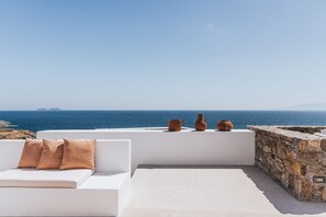 Un espacio exterior con asientos blancos, una pared de piedra, dos macetas de terracota y vistas al mar.