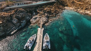 A coastal area with white buildings, a clear turquoise sea, and a bridge connecting to a dock with boats. - Cali Mykonos (Mykonos)