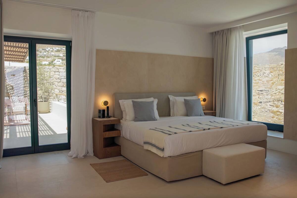 A modern bedroom with a large bed, two bedside tables with lamps, a small seating area, and a view of a mountainous landscape through the windows.