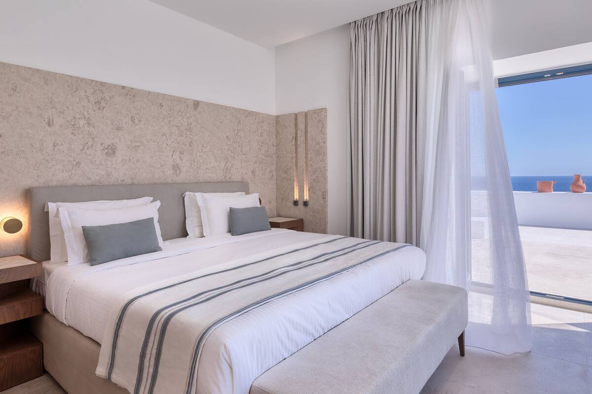 A modern bedroom with a large bed, a nightstand, and a view of the sea through sheer curtains.