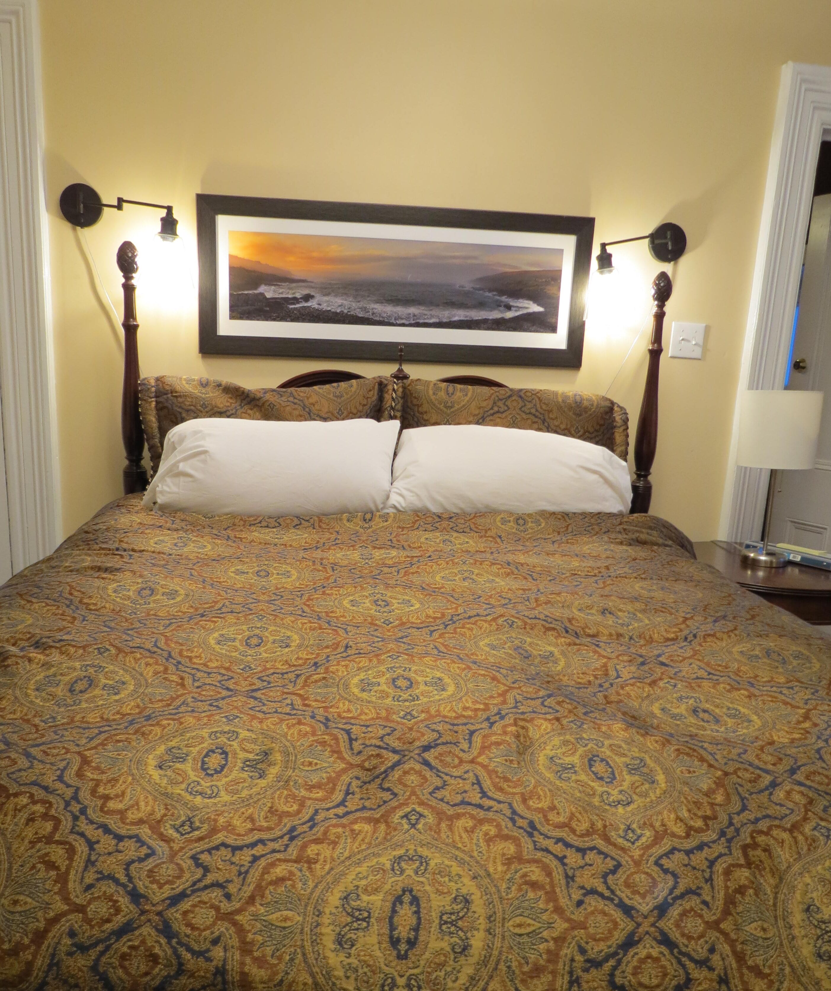 Una cama con una colcha estampada, dos almohadas blancas, un cabecero con una imagen encima y dos lámparas fijadas a la pared.