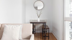 A living room with a beige sofa, a small table with two chairs, a white vase with branches, and a round mirror on the wall.