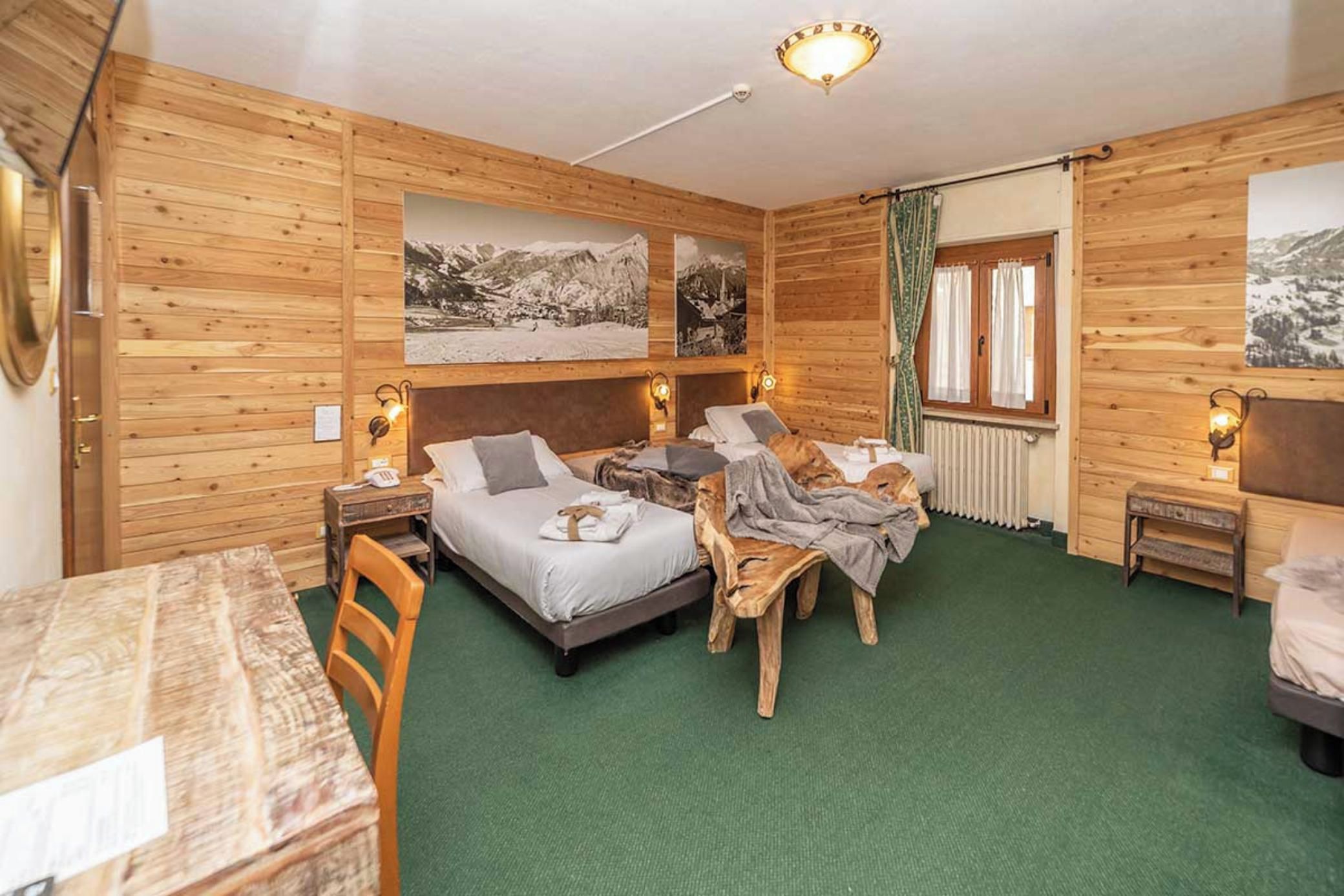 A wooden cabin room with a bed, a small table, and a chair. There are two wall-mounted lamps and a window with curtains.