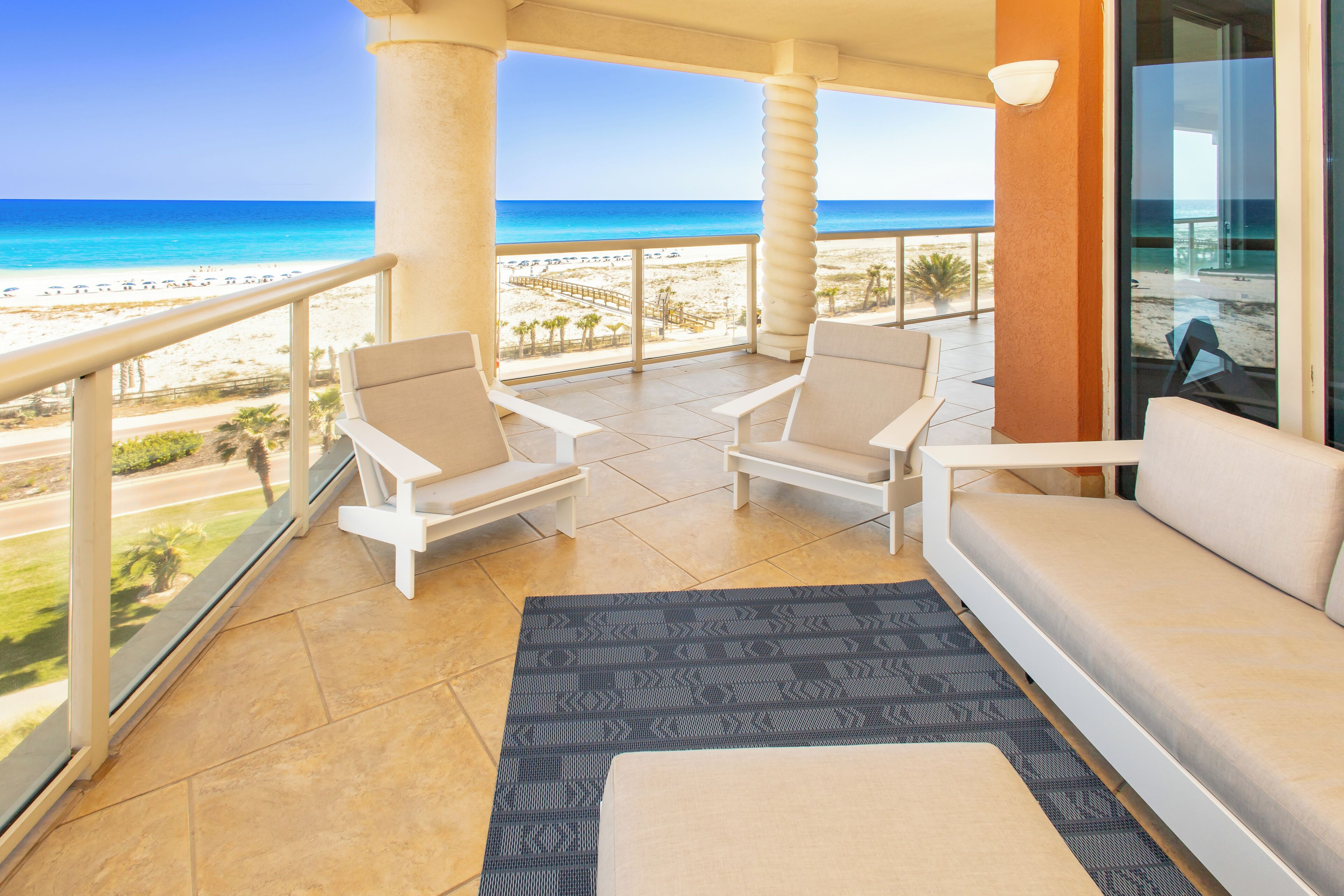 Un balcon avec vue sur la plage, deux chaises blanches, un canapé et un tapis à motifs.
