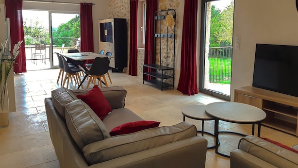 A spacious living room with a sofa, a coffee table, and a TV. There is a dining area with chairs and a table, and a balcony with a view of greenery.