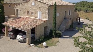A stone house with a tiled roof, a car parked in the driveway, and a staircase leading to the entrance.