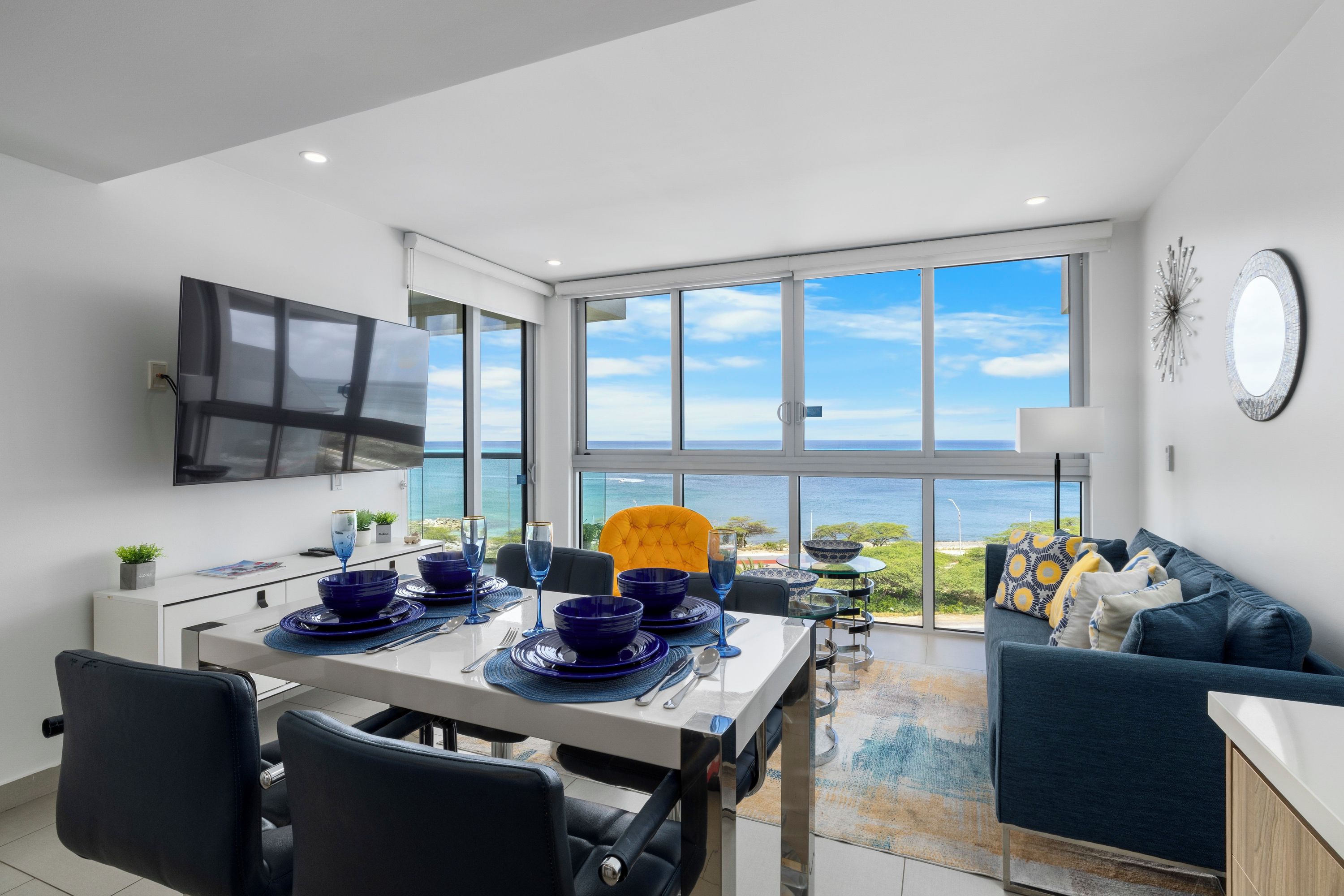 Ein moderner Essbereich mit Meerblick, einem Flachbildfernseher und einer gemütlichen Sitzecke mit Sofa und Kissen.