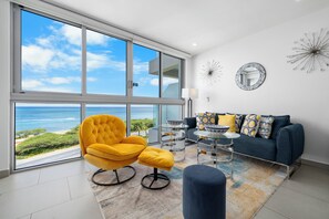 A modern living room with a large window overlooking a coastal view, a yellow armchair, a blue sofa, and decorative wall art. - Azure Beachfront Oasis: 5th Fl Ocean View & Balcony near Eagle Beach (Noord)