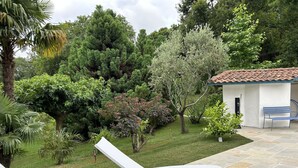 A patio with a white lounge chair, a small white building with a tiled roof, and a variety of trees and shrubs in the background. - In a green setting, very nice house . (Arcangues)