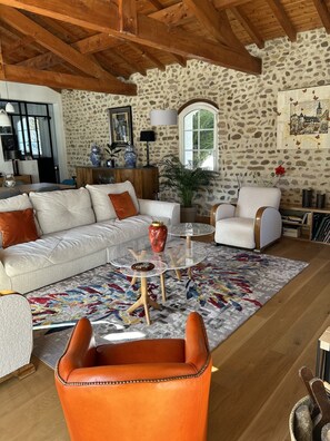 A cozy living room with a stone wall, wooden ceiling beams, a white sofa, a leather armchair, and a glass coffee table.
