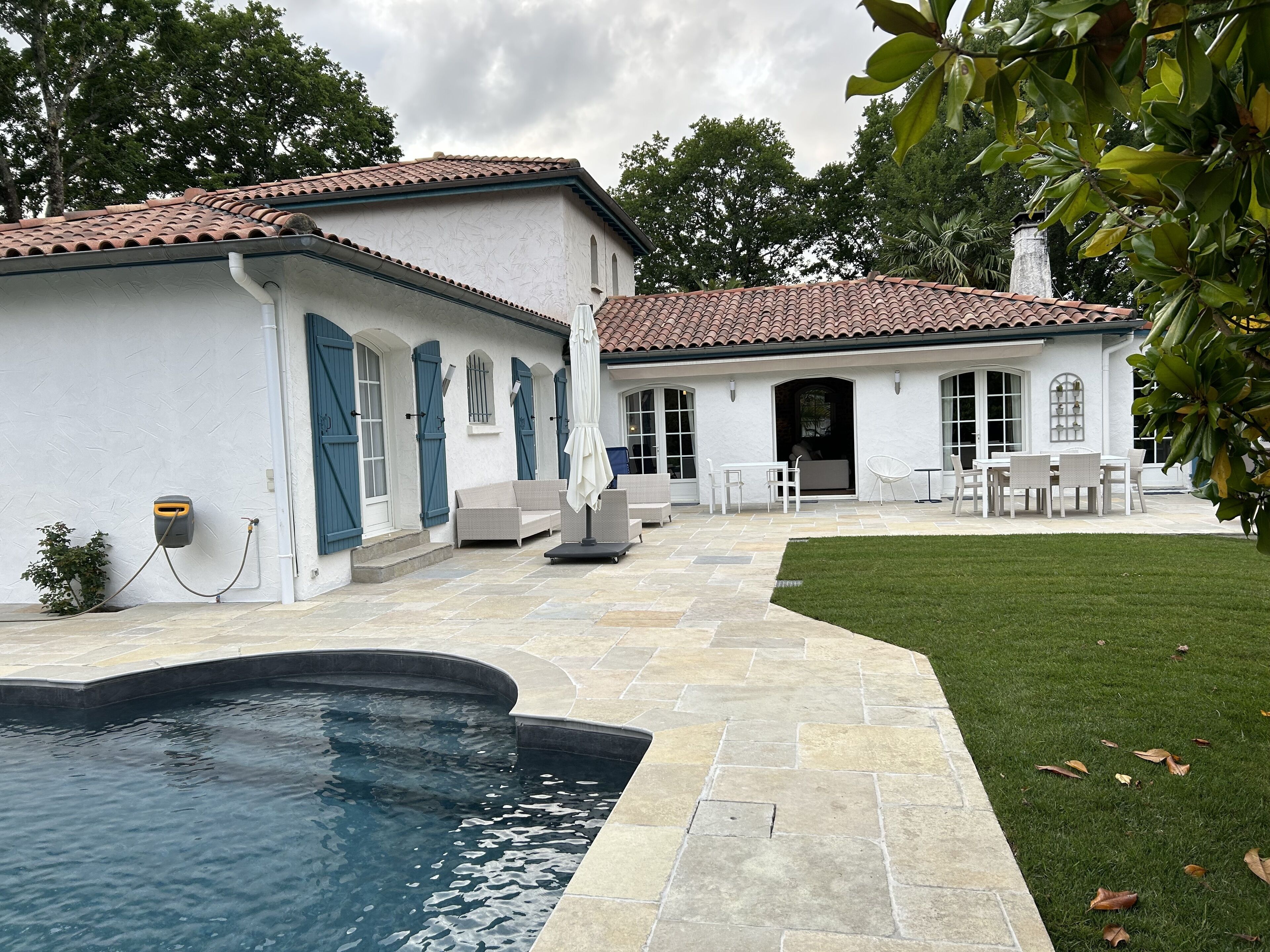 Une piscine entourée d’une terrasse en pierre, un patio couvert avec des meubles d’extérieur, et un bâtiment blanc avec des volets bleus.