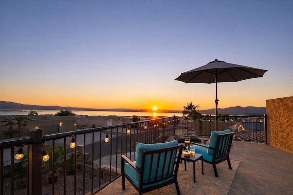 Ein Balkon mit zwei Stühlen und einem Tisch, der Blick auf einen Sonnenuntergang über Wasser und Berge bietet.