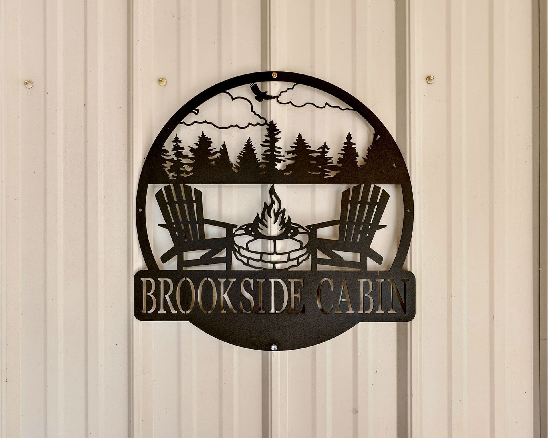 Pyöreä logo, jossa on kaksi Adirondack-tyylistä puutarhatuolia, nuotio ja puita, ja jossa lukee teksti: ”BROOKSIDE CABIN”.
