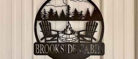 Pyöreä logo, jossa on kaksi Adirondack-tyylistä puutarhatuolia, nuotio ja puita, ja jossa lukee teksti: ”BROOKSIDE CABIN”.