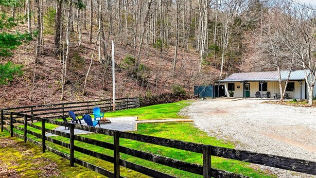 A rustic cabin with a gravel driveway, a fenced area with chairs, and a forested backdrop. - Lazy Lane Cabins - Brookside Cabin (South Bloomingville)