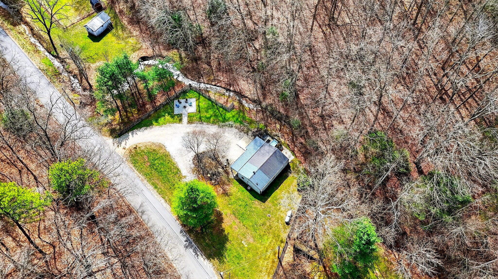An aerial view of a property with a fenced yard, a small house with a sloped roof, and a paved driveway.