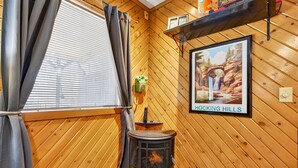 A wooden interior with a wood-burning stove, a window with curtains, and a framed picture on the wall. - Lazy Lane Cabins - Brookside Cabin (South Bloomingville)