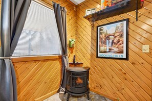 A wooden interior with a wood-burning stove, a window with curtains, and a framed picture on the wall.