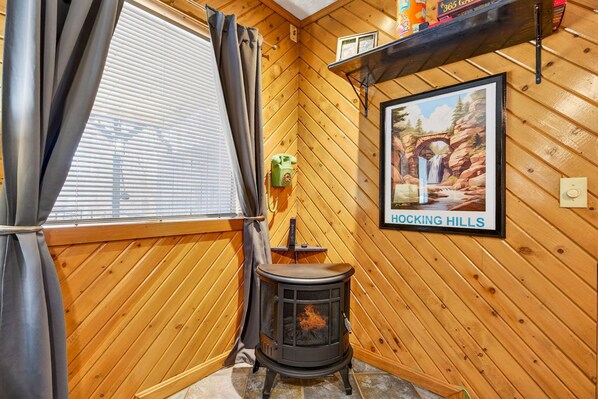 A wooden interior with a wood-burning stove, a window with curtains, and a framed picture on the wall.