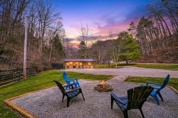 A rustic outdoor seating area with chairs and a fire pit, a small building with lights on, surrounded by trees and a gravel path.