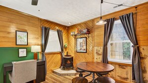 A cozy room with a wooden interior, a round table with stools, a small stove, and a desk with a chair. - Lazy Lane Cabins - Brookside Cabin (South Bloomingville)