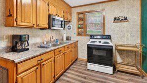 A kitchen with wooden cabinets, a microwave, a stove, a coffee maker, and a window with blinds. - Lazy Lane Cabins - Brookside Cabin (South Bloomingville)