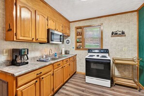 Una cocina con armarios de madera, microondas, estufa, cafetera y ventana con persianas.