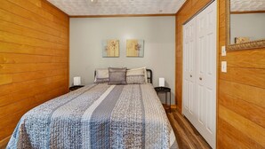 A bedroom with wooden paneling, a bed with a patterned bedspread, two wall-mounted paintings, a ceiling fan, and a white door. - Lazy Lane Cabins - Brookside Cabin (South Bloomingville)