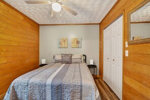 Un dormitorio con paneles de madera, una cama con colcha estampada, dos cuadros en la pared, un ventilador de techo y una puerta blanca.