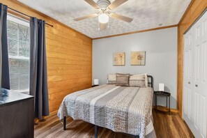 A bedroom with a wooden ceiling, a bed with a patterned bedspread, a nightstand, and a window with curtains.