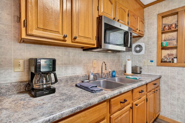Una cocina con gabinetes de madera, un microondas de acero inoxidable, una cafetera y un lavamanos.