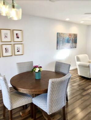 Un comedor con una mesa redonda de madera rodeada por seis sillas tapizadas de color gris. Hay un florero con flores sobre la mesa, y la habitación tiene una pared blanca con cuadros y una pintura.