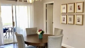 Un coin repas avec une table ronde, six chaises et un lustre. Des tableaux encadrés ornent le mur, et une porte donne accès à une autre pièce.