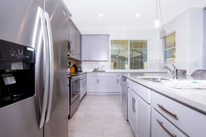 Une cuisine moderne avec des meubles blancs, des appareils en acier inoxydable et une grande fenêtre équipée de stores.