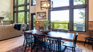 A dining area with a wooden table and chairs, large windows, and a hanging light fixture. - Luxury Smoky Chalet, Indoor Pool, Hot Tub, Theater,  Sleep 12 in 4 King Suites, Indoor Fireplace, Outdoor Fire Pit, Game Loft & Scenic Views, 20 Min to Dollywood, The Island & Parkway, Dog-Friendly, 24 Hr Hospitality (Sevierville)