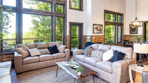 A spacious living room with large windows, two sofas, a coffee table, and a lamp. - Luxury Smoky Chalet, Indoor Pool, Hot Tub, Theater,  Sleep 12 in 4 King Suites, Indoor Fireplace, Outdoor Fire Pit, Game Loft & Scenic Views, 20 Min to Dollywood, The Island & Parkway, Dog-Friendly, 24 Hr Hospitality (Sevierville)