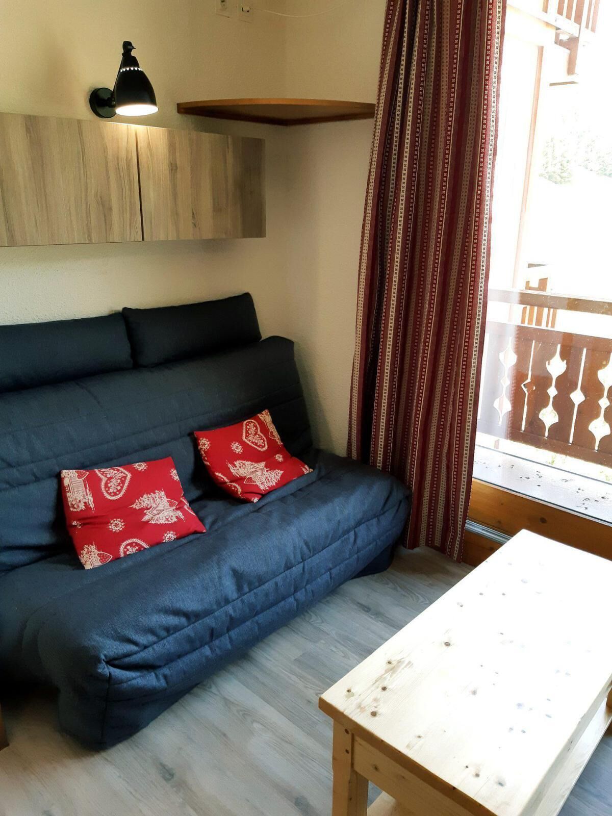 Une pièce avec un canapé bleu foncé, deux coussins rouges à motifs, une petite table en bois, un luminaire fixé au mur et une fenêtre ornée de rideaux à motifs.