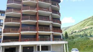 A multi-story building with balconies, a parking area, and a hilly landscape in the background. - 100m from resort center, 4th floor, view mountain, balcony, parking, tv, ski locker, 40m² (Les Deux Alpes)