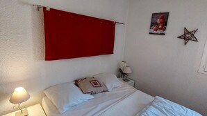 A bedroom with a bed, a nightstand, a lamp, a red curtain, and two wall decorations. - 100m from resort center, 4th floor, view mountain, balcony, parking, tv, ski locker, 40m² (Les Deux Alpes)